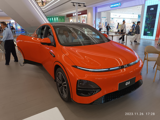 Ein oranges Elektroauto in einer Ausstellungshalle, umgeben von Menschen, mit Stühlen, einem Wagen, Geschäften, Namensschildern und Lichtern im Hintergrund.