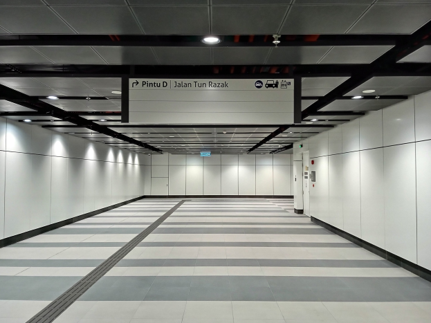 Langer Flur mit weißen Wänden, schwarzem und weißem Schachbrettboden, Deckenleuchten und einer Tafel mit Text oben, der an eine Tiefgarage erinnert.