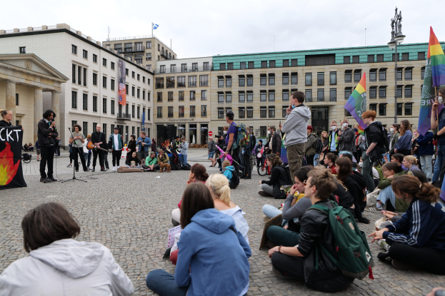 Eine Gruppe von Menschen sitzt auf dem Boden vor einer Menge mit Fahnen und Schildern, mit einer Person, die in ein Mikrofon spricht, einer Statue und Gebäuden im Hintergrund während einer anti-schwulen Demonstration in Berlin.