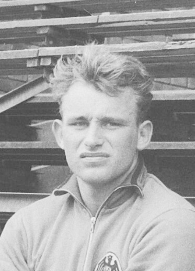 Schwarzes und weißes Foto eines Mannes in einer Jacke, der vor einem Stapel Holzplanken steht, der als legendärer deutscher Fußballspieler gilt.