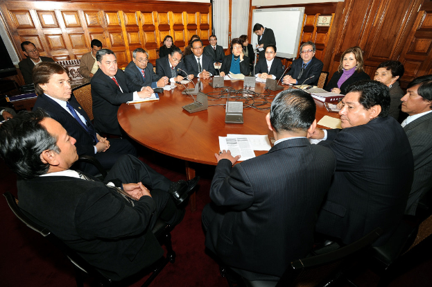Eine Runde-Tisch-Besprechung mit Delegierten, die Ã¼ber Mikrofone, Papiere, Kabel und Dateien auf dem Tisch diskutieren, mit einer hÃ¶lzernen Wand im Hintergrund.