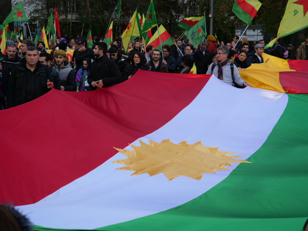 Eine große Gruppe von Menschen mit Fahnen vor einem Gebäude, mit Bäumen und Pfählen im Hintergrund, wahrscheinlich an einer Protestaktion in Kurdistan teilnehmend.