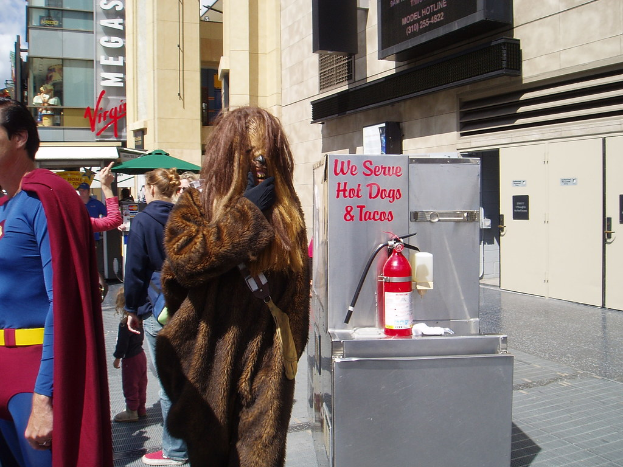 Eine Menschenmenge, einige in Kost√ľmen, versammelte sich vor Geb√§uden mit Schildern, einer Box mit Schrift und einem Feuerl√∂scher.