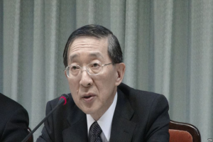 Ein Mann in Anzug und Krawatte sitzt an einem Tisch mit einem Mikrofon, trägt eine Brille, in einem formellen Setting mit einem Vorhang im Hintergrund, während die Hand einer anderen Person auf der linken Seite des Bildes sichtbar ist.