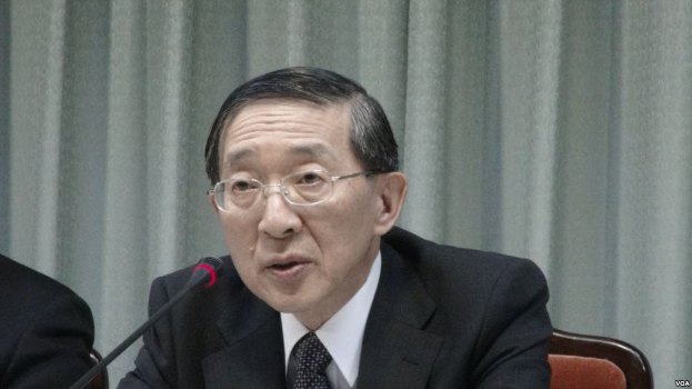 Ein Mann in Anzug und Krawatte sitzt an einem Tisch mit einem Mikrofon, trägt eine Brille, in einem formellen Setting mit einem Vorhang im Hintergrund, während die Hand einer anderen Person auf der linken Seite des Bildes sichtbar ist.