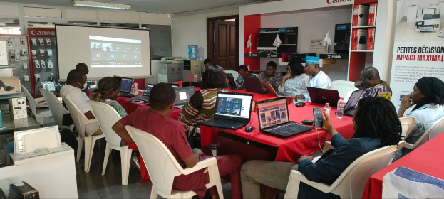 Eine Gruppe von Menschen sitzt um einen Tisch mit Laptops, Mäusen, Flaschen und anderen Gegenständen und nimmt an einem Workshop zum digitalen Marketing teil, mit einem Banner, einem Display, einer Tür, Deckenlampen und elektronischen Geräten im Hintergrund.