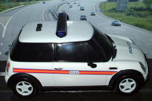 Ein weißer Polizeiwagen steht im Vordergrund, während viele Autos auf der Straße dahinter fahren, umgeben von Gras und Bäumen auf beiden Seiten, in einer ländlichen Umgebung.