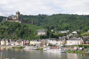 Eine malerische Aussicht auf den Rhein in Deutschland mit einer Burg auf einem Hügel, Booten auf dem Fluss, Fahrzeugen auf einer nahen Straße und einem bewölkten Himmel.