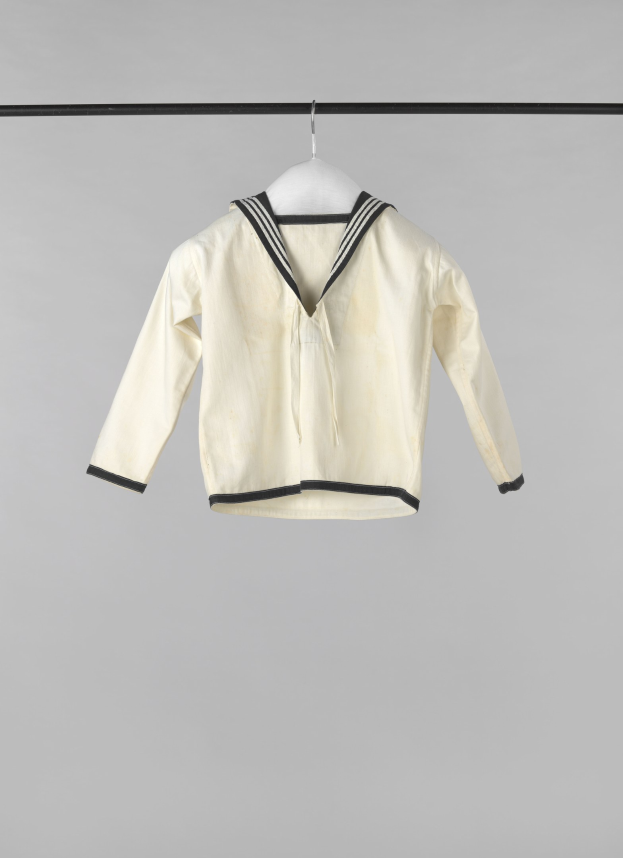 Eine weiße Matrosenjacke mit Kragen, langen Ärmeln und Knopfleiste hängt an einer Wäscheleine vor einem weißen Hintergrund, aus leichtem, geripptem Stoff.