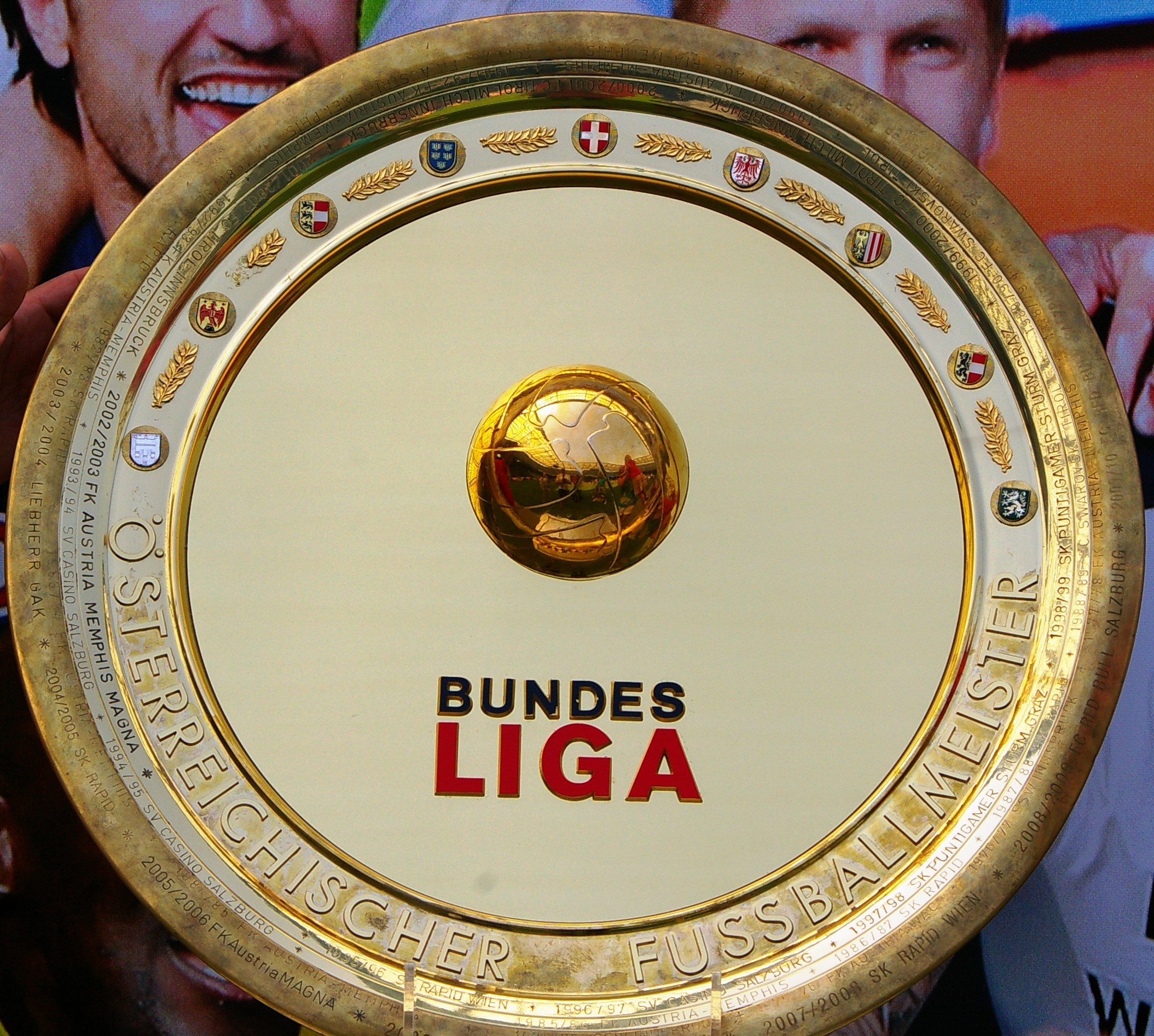 Goldener Schild mit "Bundesliga"-Text, gehalten von einem Mann, mit einem Banner im Hintergrund, das drei Personen zeigt.