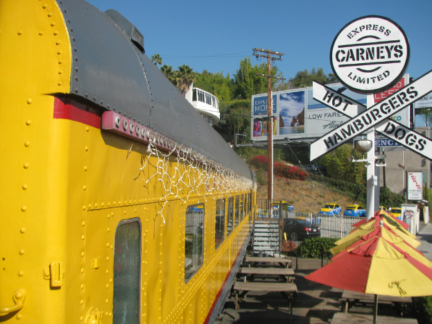 Eine mobile Kantine ist neben Bänken und Stufen geparkt, mit Schirmen, Pflanzen und einem Geländer in der Nähe. Fahrzeuge, Pfosten, Bretter und Werbetafeln sind im Hintergrund zu sehen, sowie ein Gebäude, Bäume und den Himmel.