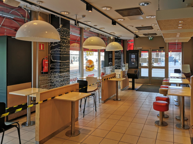 Innenraum eines Fast-Food-Restaurants mit Tischen, Stühlen, Hockern, einem Boden, Deckenleuchten, einer wandmontierten Feuerlöschanlage, Glastüren und weiterer Einrichtung.