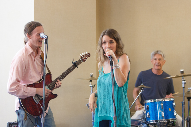 Ein Mann spielt Gitarre und lächelt, eine Frau in einem blauen Kleid singt in ein Mikrofon, und eine Person sitzt an einem Tisch und hält Drumsticks vor Musikinstrumenten, mit einem Mikrofonständer und einer cremefarbenen Wand im Hintergrund.