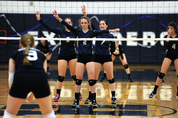 Drei Mädchen auf einem Volleyballfeld mit erhobenen Händen vor einem Netz und einem Zaun im Hintergrund.