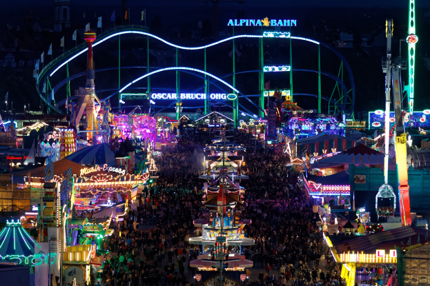 Eine große Menschenmenge versammelt sich um einen hell erleuchteten Karneval in der Nacht, umgeben von Vergnügungsgeräten, Pfosten, Flaggen und Namensschildern, mit einem dunklen Hintergrund.