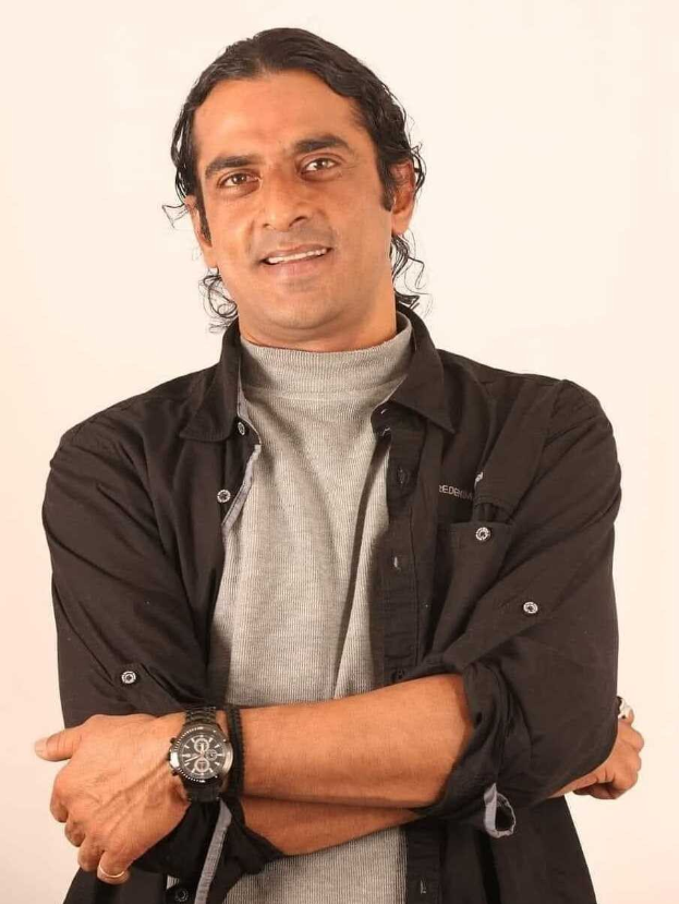 Ein Mann in einem schwarzen und aschfarbenen Kleid mit überschlagenen Armen, der eine Uhr tr├Ągt und gegen einen wei├čen Hintergrund steht, der scheinbar in Gedanken versunken ist.