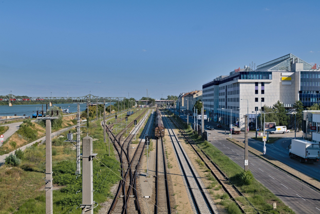Ein Zug fährt neben einer Stadtlandschaft mit Gebäuden, Infrastruktur, Fahrzeugen, Grünflächen und einer Brücke unter einem bewölkten Himmel.