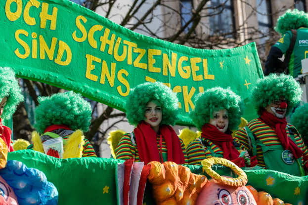 Eine Gruppe von Clowns in bunten Kostümen marschiert durch eine Straße, eine Fahne mit Text haltend, mit Bäumen und einem Gebäude im Hintergrund.