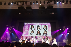 Eine Gruppe von Menschen, die auf einer hell erleuchteten Bühne performen, wobei einer ein Mikrofon hält und ein anderer ein Instrument spielt, während die Menge mit Kameras unten und ein großer Bildschirm Bilder von BK48 zeigt.