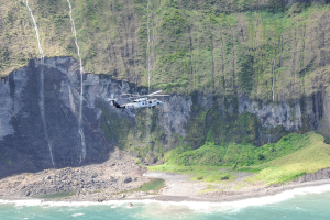 Ein Hubschrauber fliegt über das Meer neben einer Klippe, mit Wasser darunter, Felsen und Gras am Boden und Bäumen auf dem Berg im Hintergrund.
