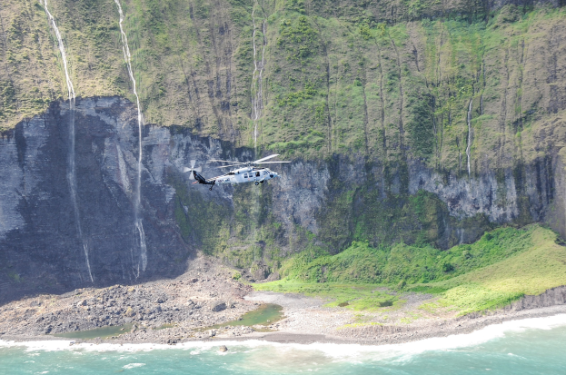 Ein Hubschrauber fliegt über das Meer neben einer Klippe, mit Wasser darunter, Felsen und Gras am Boden und Bäumen auf dem Berg im Hintergrund.