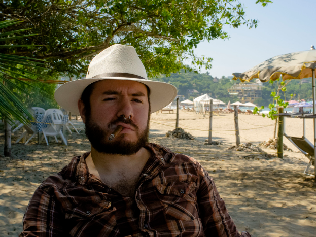 Ein Mann in Hemd und Hut raucht eine Zigarre im Freien mit Stühlen, Bäumen, Zelten, Häusern, einem Schirm, Pfählen und Felsen im Hintergrund.