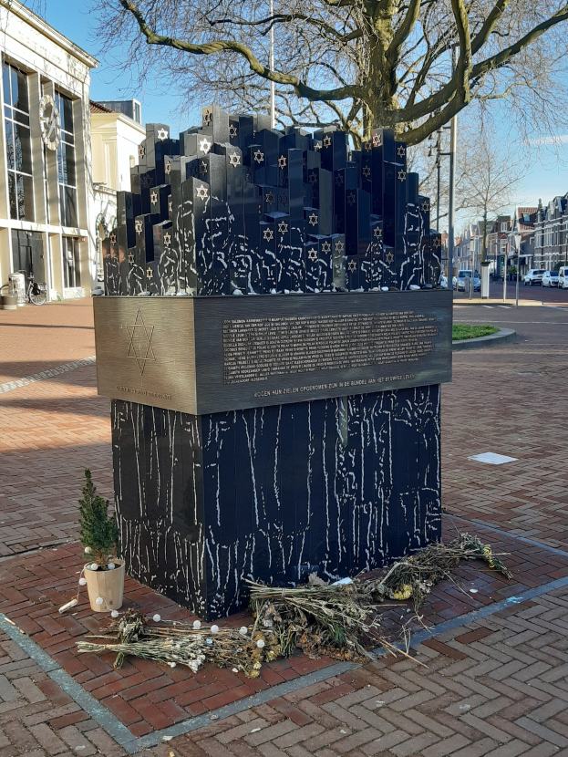 Denkmal in einem Stadtplatz für Holocaust-Opfer, mit einem Baum davor, einem Topf auf dem Boden, verstreuten trockenen Blättern und Gebäuden, Fahrzeugen, Masten und Bäumen im Hintergrund.