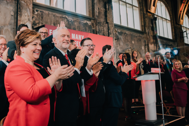 Eine Gruppe von Menschen, wahrscheinlich Liberale, steht vor einer Menge und klatscht feierlich, mit einem Podium, Mikrofon und Texttafel rechts und Stühlen, Banner, Wand, Fenstern und Lichtern im Hintergrund.