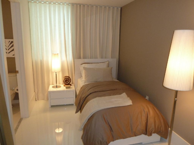 Ein Zimmer mit einem Bett, zwei Kissen, einem Nachttisch mit verschiedenen Gegenständen, einem Handtuch, einer Rolle Papier, einem Vorhang und zwei Lampen, mit einem Badezimmer im Hintergrund.