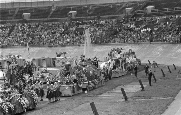 Schwarzes und weißes Foto eines Umzugs in einem Stadion mit Menschen, die stehen und sitzen, einem zentralen Springbrunnen und Blumengebinden auf den Fahrzeugen.