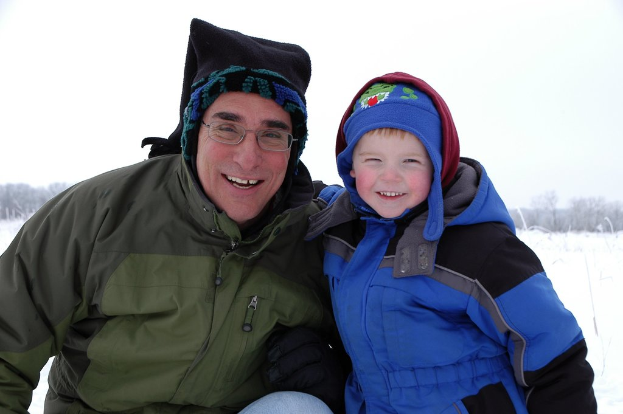 Ein Kind in einer blauen Jacke steht rechts lachend, während eine sitzende Person links lächelt, mit einer verschneiten Landschaft, Bäumen und Himmel im Hintergrund.