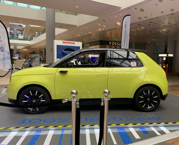 Gelbes wasserstoffbetriebenes Elektroauto vor einem Gebäude mit Absperrpollern, Seilen, Bannern, einer Person im Auto, einer Säule, stehenden Menschen, Schildern, einer Topfpflanze, Deckenleuchten und Lüftern.