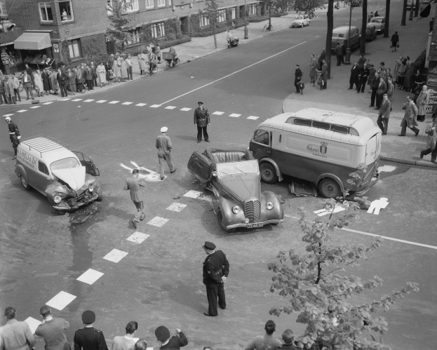 Schwarz-weißes Bild einer Autounfall-Szene an einer Straßenkreuzung mit mehreren Fahrzeugen, darunter ein Lieferwagen, und Zuschauern; Gebäude, Bäume, Pfosten und Texttafeln im Hintergrund.