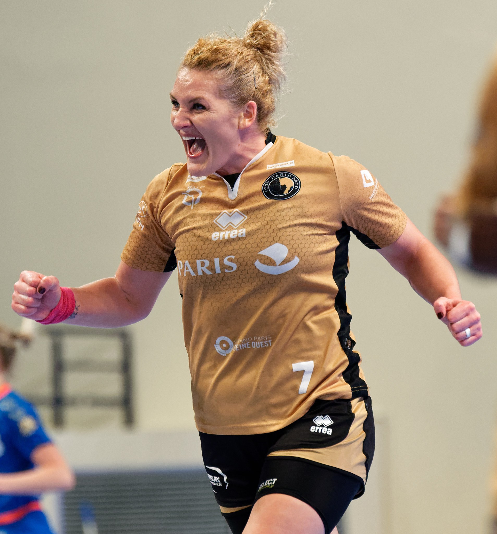 Eine Frau in braunem T-Shirt und schwarzen Shorts rennt auf einem Futsalplatz, feiernd mit triumphierendem Gesichtsausdruck, während zwei andere Frauen im Hintergrund unscharf sind.