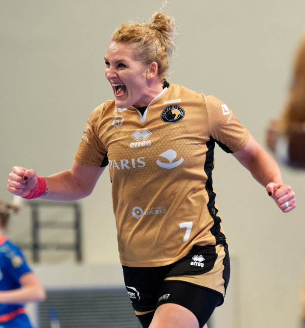 Eine Frau in braunem T-Shirt und schwarzen Shorts rennt auf einem Futsalplatz, feiernd mit triumphierendem Gesichtsausdruck, während zwei andere Frauen im Hintergrund unscharf sind.