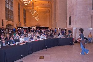 Eine Konzertszene mit einer Menschenmenge, einem Mann, der rechts Gitarre spielt und singt, einer Wand und Fenstern im Hintergrund, Lampen oben und einem Boden unten.
