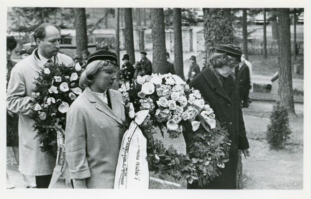 Eine Gruppe von Menschen hölt Blumensträuße und steht zusammen bei einer Gedenkfeier, mit Bäumen und Gebäuden im Hintergrund, dargestellt in Schwarz-Weiß.