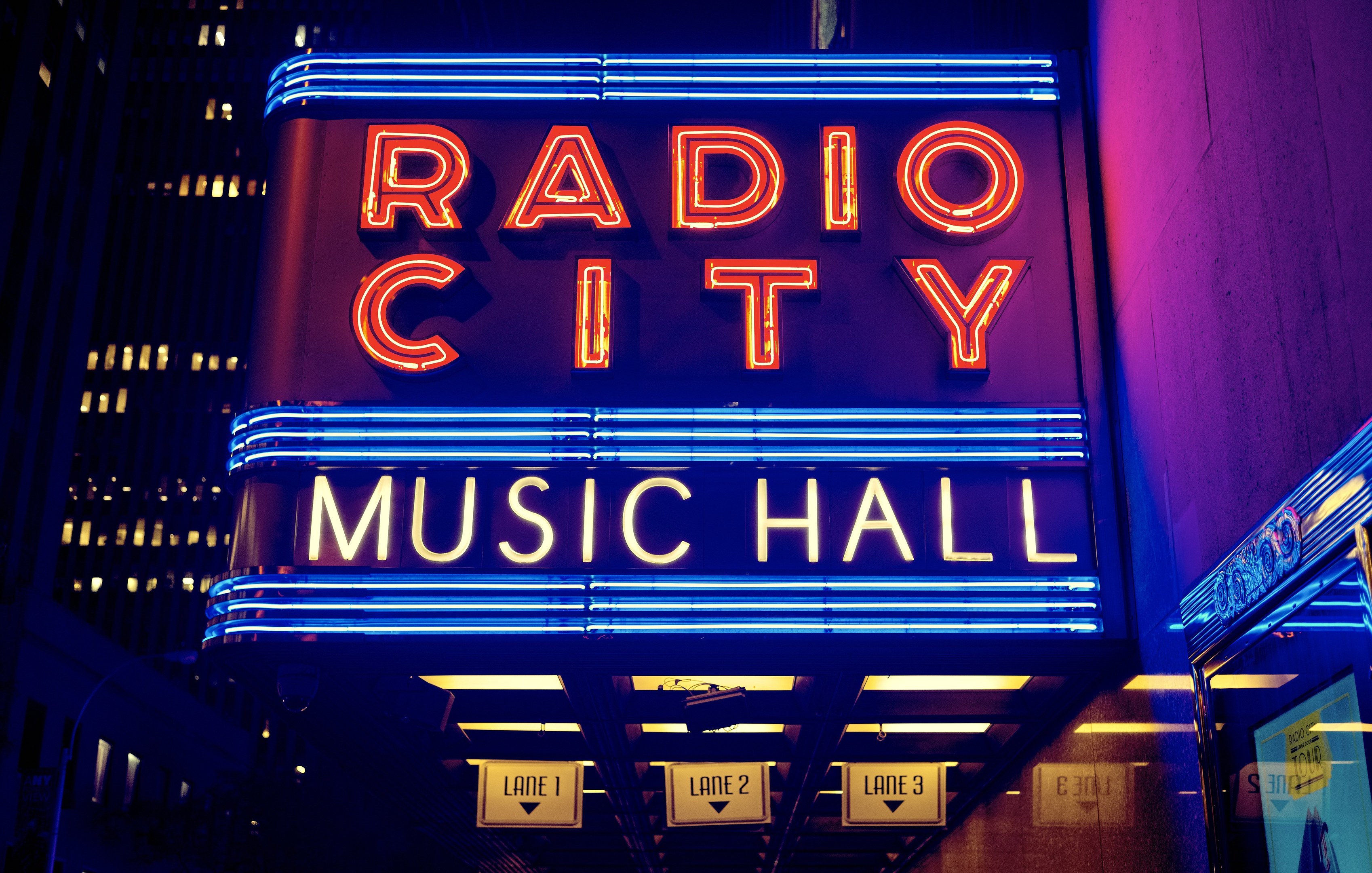 Außensicht des Radio City Music Hall in New York City bei Nacht, hell erleuchtet mit hohen Gebäuden im Hintergrund, mit einer 'Radio City'-Leuchtschrift und mehreren kleineren Schildern auf der rechten Seite.