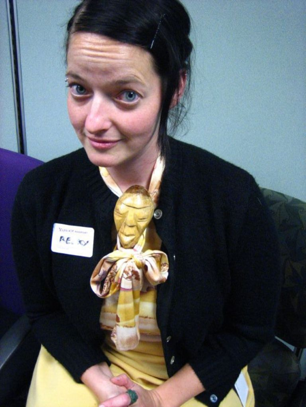 Eine Frau sitzt auf einem Stuhl und trägt eine schwarze Jacke und ein gelbes Kleid, mit einem Abzeichen an ihrer Kleidung, vor einer Wand.