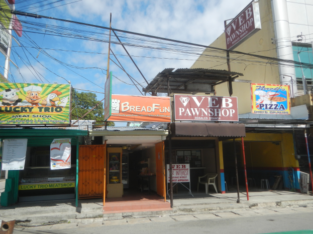 Eine belebte Straßenszene mit einer Bäckerei in der Mitte, die verschiedene Lebensmittel anbietet, umgeben von Gebäuden, Fahrzeugen und Bäumen unter einem bewölkten Himmel.