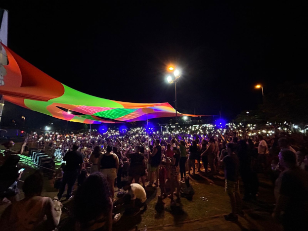 Eine große Menschenmenge steht vor einem Zelt in der Nacht, beleuchtet von bunten Lichtern und umgeben von beleuchteten Pfählen, mit einem Gebäude auf der linken Seite und dem Nachthimmel im Hintergrund.