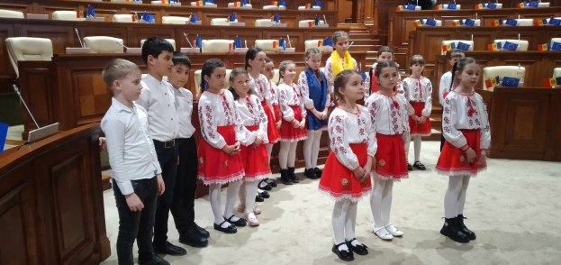 Eine Gruppe von Kindern in weißen und roten Kleidern steht in einem Raum mit einem Podium, Mikrofon, Stühlen, Tischen und Gegenständen im Hintergrund, tragend Flaggen als Teil einer Nationalversammlung.
