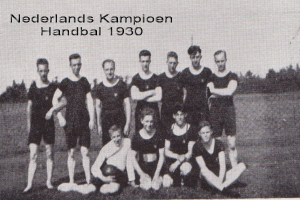 Ein Schwarz-Weiß-Foto einer Handballmannschaft aus dem Jahr 1930, mit einigen Mitgliedern im Vordergrund stehend und sitzend, Bäumen und einem klaren Himmel im Hintergrund und Text oben auf dem Bild.
