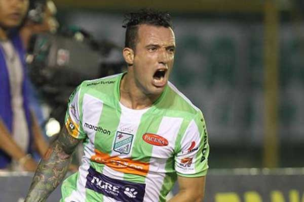 Ein Fussballer in grüner und weisser Uniform mit Tattoos auf dem Arm feiert nach einem Tor, mit unscharfen Hintergrundfiguren und einer Tafel.