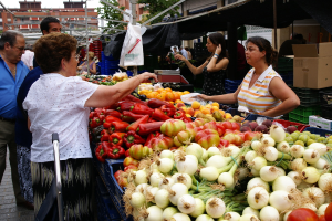 Eine Gruppe von Menschen steht vor Gebäuden und Bäumen, mit Gemüse in Plastikbehältern und einer Kartonbox in der Nähe.