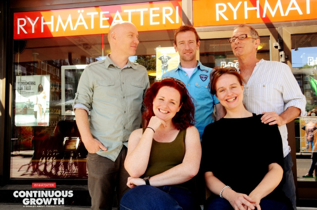 Drei Männer stehen und zwei Frauen sitzen vor ihnen, alle lächeln, mit Geschäften im Hintergrund, die Brillengläser anbieten, und einem Schild mit der Aufschrift "RHYME TEATTERI" über einem der Geschäfte.