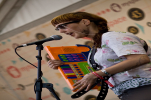 Eine Frau in einem weißen Hemd und Sonnenbrille singt in ein Mikrofon und spielt Klavier, mit einem Plakat an der Wand hinter ihr.