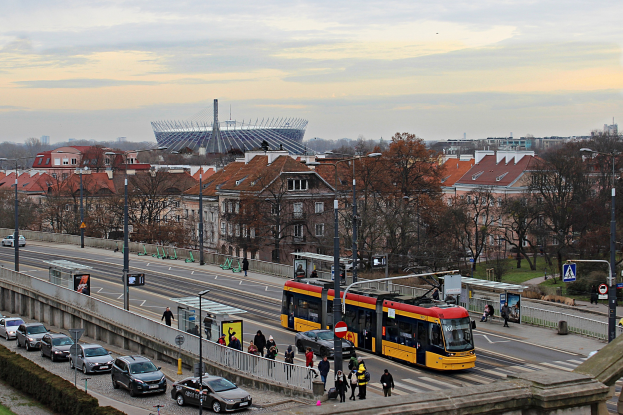 Eine gelbe und schwarze Straßenbahn fährt auf einer von hohen Gebäuden gesäumten Stadtstraße, umgeben von Autos, Fußgängern, Bäumen und Laternen unter einem bewölkten Himmel.