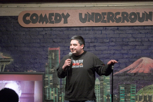 Ein Mann steht mit einem Mikrofon in der Hand und spricht, mit einem Bild an der Wand im Hintergrund, einem Fernseher und Text an der Wand.