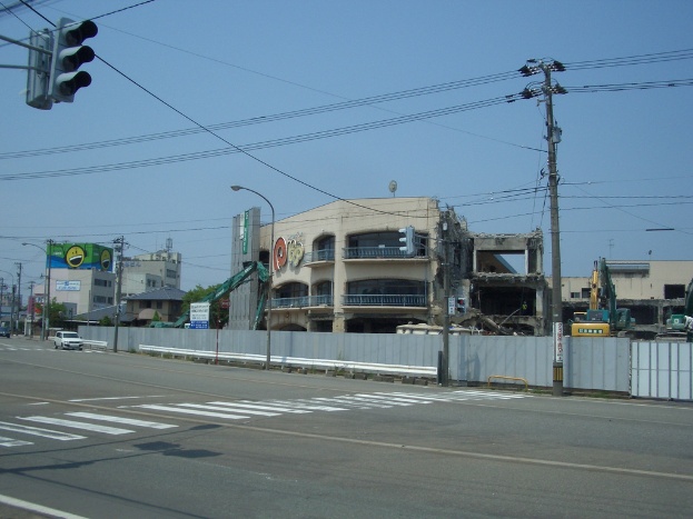 Eine Stadtstraße mit Fahrzeugen, Strommasten, Laternen und einer Ampel auf der linken Seite, Gebäuden, Bäumen und dem Himmel im Hintergrund und einem abbruchreifen Gebäude im Vordergrund.